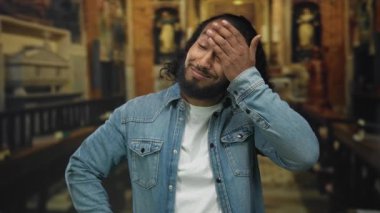 Bir kilise binasında gözünü ovuşturan bir adam, eli kot ceket ve beyaz gömlek ile kaplı; yorgunluk yansıması.