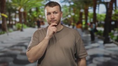 Elleri palmiye ağacında çene üstüne uzanan adam sokak gezintisi, sıradan tişört ve sakal, dışarıda beklerken ileriye bakıyor; düşünceli..