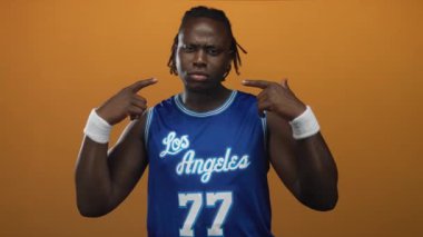 Man points finger to face in studio with orange backdrop, wearing los angeles basketball jersey 77 and white wristbands; confidence.