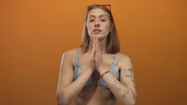 Dövmeli ve bikinili bir kadın izole edilmiş turuncu bir arka plana karşı kırmızı saç tokası ve güneş gözlükleriyle duygularını ifade ediyor..