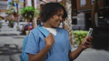 Mavi önlüklü gülümseyen genç kadın sırtında sırt çantası ve parşömen taşır. Şehir caddesinde akıllı telefon; mutluluk..