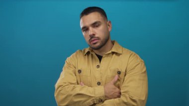 İspanyol adam canlı mavi stüdyo ortamında kollarını çaprazlıyor. Hardal rengi ceket giyiyor. Yanlara bakıyor..