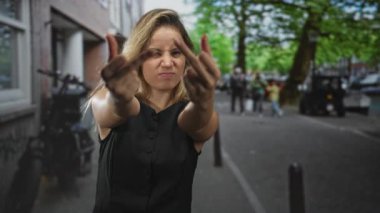 Genç sarışın kadın, şehir caddesinde iki orta parmağını da kameraya gösteriyor. Yüzünü sımsıkı kavrıyor ve ellerini uzatıyor..