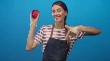 Önlük ve tek kullanımlık eldiven giyen kadın kırmızı elmayı tutuyor ve parmağıyla mavi stüdyoda meyveyi işaret ediyor, çizgili gömlek görünür;.
