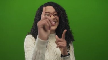 Kıvırcık saçlı, kırmızı gözlüklü, gülümseyen ve kameraya işaret eden, yeşil stüdyoda beyaz kazak giyen, kendine güvenen bir kadın..