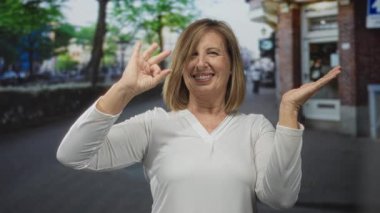 Gülümseyen orta yaşlı beyaz kadın, gün içinde canlı şehir caddesinde el ve el işaretleri ve açık el hareketleriyle tamam diyor..