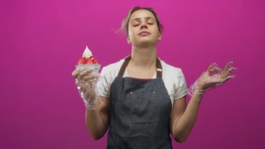 Çilekli bir tatlı bardağını çırpılmış kremayla tutan ve eflatun arka planlı bir stüdyoda el hareketi yapan bir kadın..