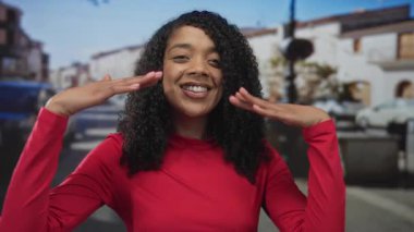 Bir şehir caddesinde elleri kırmızı uzun kollu bir bluzla gülümseyen ve çerçeveleten bir kadın..