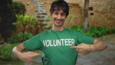 Adam gülümsüyor ve parkın dışındaki yeşil güneşli parkta gönüllü gömleği işaret ediyor..