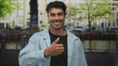 Young hispanic man in casual jacket and black shirt gives thumbs up on street by canal and parked bicycle rack; approval.