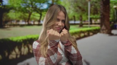 Kadın, sarı saçlı, yeşil güneşli parkta çıplak yumruğunu öne doğru sallıyor..