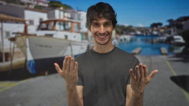 Adam mavi gökyüzü altında beyaz teknelerin yanında güneşli sokak rıhtımında her iki avuç içi jestini gösterir; sıcak davet.