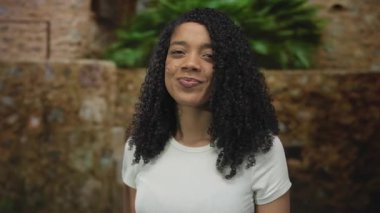 Kıvırcık saçlı, gülümseyen ve doğrudan kameraya bakan yeşil yapraklı bir kadın..