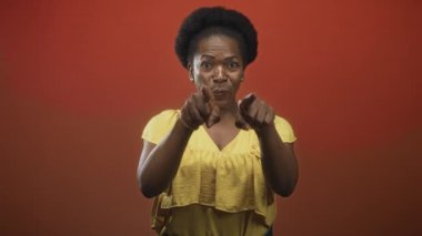 İki işaret parmağı önde olan kadın, ağzı açık ve elleri stüdyoda görünür durumda, turuncu arka plan; sürpriz bir güven..