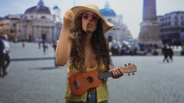 Ukulele 'yi elinde tutan kadın tarihi bir binanın önünde geniş bir şapka ve yuvarlak güneş gözlüğü takıyor..