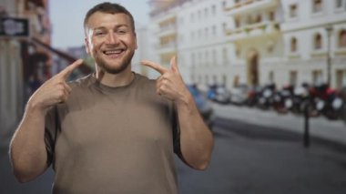 Genç beyaz adam gülümsemesini işaret ediyor dişlerini gösteriyor ve işlek bir şehir caddesinde kahverengi bir tişört giyiyor..