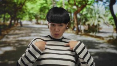 Yeşil bir parkta açık havada duran çizgili kazaklı kısa saçlı kadın güneşli bir günde gür ağaçların altında kendini komik bir ifadeyle gösteriyor..