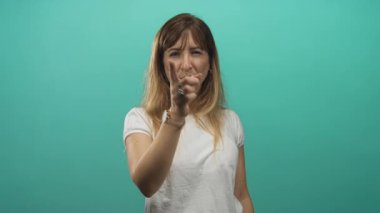 Genç bir kadın parmağıyla kamerayı işaret ederek yüzük takıyor ve beyaz t-shirtlü Teal stüdyosunun arka planında izliyor..