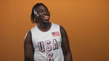 Portakal rengi stüdyoda Amerikan bayraklı Amerikan basketbol forması giyen ve ağzı açık gülümseyen bir adam..