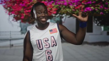 ABD basketbol formalı genç adam gülümsüyor ve çıplak kol ve mavi bileklik ile barış işareti tutuyor ağaçlar inşa etmek ve çiçek açmak dışında; neşe vatanseverliği.