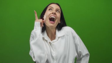Genç İspanyol kadın enerjik bir şekilde gülümsüyor beyaz bir tişörtle izole edilmiş yeşil bir arka plan duvarına karşı, şakacı bir şekilde yukarı doğru hareket ediyor ve neşe ifade ediyor.