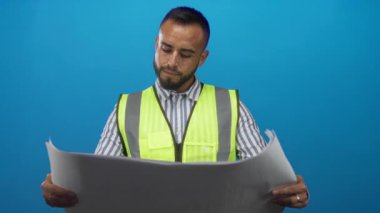 Yüksek görüş açısına sahip yelek mühendisi elinde planlar ve okuma planları olan parlak mavi arka planı olan bir stüdyoda..