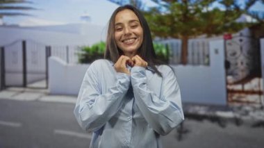 Güneşli bir caddede beyaz kapılı bir binanın önünde elleri ve ağacı olan genç bir İspanyol kadın kalp oluşturuyor..