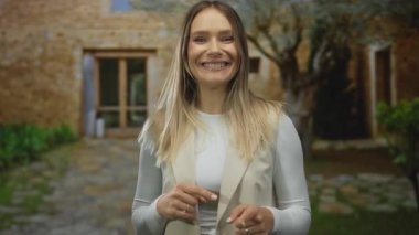 Bir kadın gülerek şehir caddesinde gün ışığında taş binalarla kamerayı işaret ediyor..