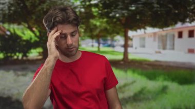 Elleri yüzü üzerinde olan bir adam yeşil bir parkta, ağaçlar ve çimlerle dolu bir binanın yanında alnını ovalıyor. Yorgunluk tefekkür ediyor..