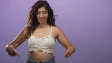 Genç İspanyol kadın çıplak göğsü işaret ediyor ve mor stüdyo duvarına bakarken gülümsüyor..