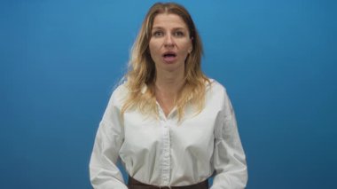 Ağzı açık, elleri havada, beyaz gömlek ve yakası açık bir kadın, sağlam mavi bir stüdyo duvarına çerçevelenmiş; sürpriz konuşma reaksiyonu.