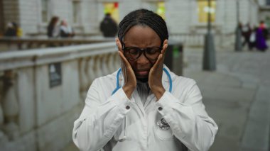Beyaz önlük ve steteskop giyen kadın doktor, şehir sokaklarında stresli görünüyor, eller gözlüklü, kentsel ortamlarda tıbbi stresi gösteriyor..