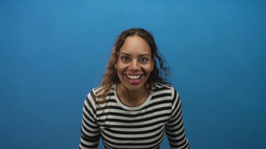 Elleri ve ağzı açık gülümseyen genç bir kadın, saçları açık, yüzü mavi stüdyoda öne dönük; sevinç kutlaması enerjisi.