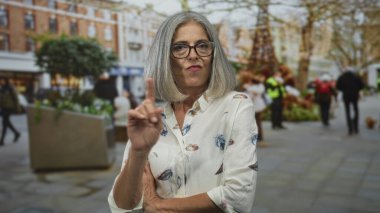 Şehir caddesinde, gün ışığında parmakla işaret eden ve çapraz silah tutan bir kadın, gri saç ve gözlük görünür; kesinlikle onaylanmamış..