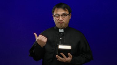 Elinde incili tutan rahip mavi arka planda düşünceli bir ifade sergiliyor.