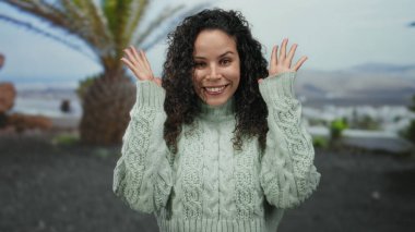 Parkta gülümseyen bir kadın palmiye ağaçlarının altında rahat bir süveter giyiyor, gündüz doğal ortamında neşeli bir ifade sergiliyor..