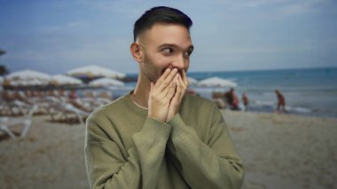 Şaşırmış ifadeye sahip genç bir adam kumlu bir sahilde elleri ve arka planda şemsiyeleri olan ağzını kapatarak güneşli bir sahil gününde beklenmedik bir anı yakalıyor..