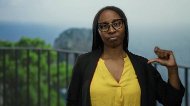 Gözlüklü ve sarı gömlekli bir kadın deniz ve yeşillik zeminiyle dışarıda duruyor, elini kaldırmış, güneşli bir günde kendine güveni ve tarzını somutlaştırıyor..