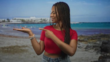 Kadın, kumsalda avuç içini göstererek kırmızı kırbaç bluzu ve bilezikler takıyor, gülümsüyor ve eline bakıyor; neşeli bir davet..