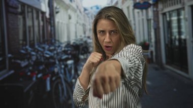 Sarışın kadın dar şehir sokağında kaldırıma parketmiş bisikletli kameraya yumruk atıyor..