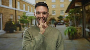 Genç adam neşeli bir şekilde gülümsüyor. Canlı bir şehir caddesinde parmaklarıyla. Açık havada genç ve samimi bir enerji ve şehir yaşamını yakalıyor..