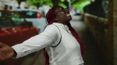 Güneşli bir günde, canlı bir şehir caddesinde pembe saçlı gülümseyen bir kadın. Beyaz bir kıyafet ve gözlük takıyor. Arka planda bulanık arabalar ve yeşil ağaçlarla çevrili..