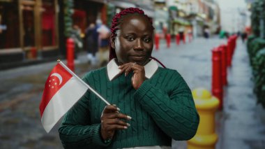 Kadın şehir caddesinde elinde Singapur bayrağı, yeşil kazak giyiyor ve gülümsüyor, şehir kültürünü ve çeşitliliğini gösteriyor..