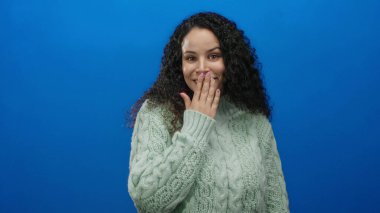 Parlak mavi arka planda yeşil kazak giyen ve kıvırcık saçlı, ağzı açık gülümseyen bir kadın izole stüdyo ortamında sürpriz ve haz dolu bir ifade veriyor.