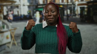 Sokakta gülümseyen, yumrukları havada, yeşil süveter ve kırmızı örgü takan bir kadın açık hava ortamında kendine güven ve güç gösterisi yapıyor..
