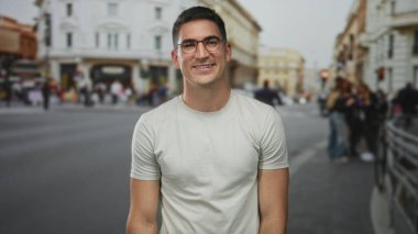 Gözlüklü bir adam gülümsüyor ve yukarı bakıyor, çıplak kolları görünür, işlek bir caddede tarihi binalarla dolu beyaz bir tişört giyiyor;.