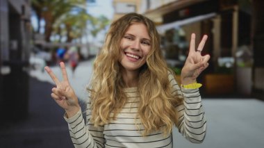 Kadın canlı bir sokakta barış işareti göstererek elini kaldırıyor, genç sarışın gülüşü, çifte jesti ve gelişigüzel tavırlarıyla..