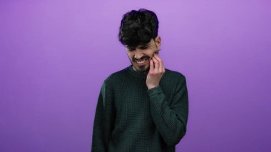 Dişi ağrılı bir adam, mor bir arka plana karşı, yeşil bir kazak giyiyor, rahatsızlık ve acı gösteriyor, izole edilmiş ve bir portre için poz veriyor..