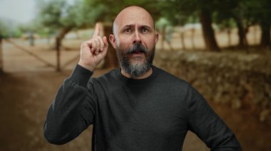 Kel sakallı bir adam, etrafı bulanık ağaçlarla çevrili bir açık hava parkında düşünceli bir şekilde el kol hareketi yaparak doğal ve sakin bir ortamda tefekküre dalmış..