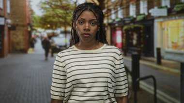 Çizgili kazaklı kadın caddede kollarını kavuşturmuş, gözleri kapalı ve kaşlarını çatmış, burun halkası görünür, düş kırıklığı yalnızlık yansıması.